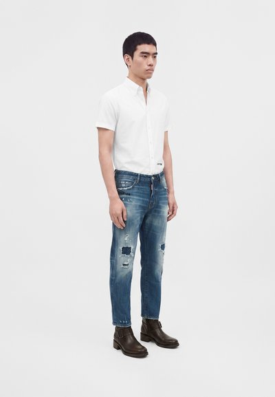 Jeune homme debout de profil portant une chemise blanche à manches courtes, un jean bleu déchiré et des bottines marron foncé.