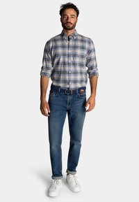 Geruite button-up shirt in blauw en beige, gecombineerd met donkere spijkerbroek en witte sneakers; heeft een opgerolde manchetten en een geweven riem.
