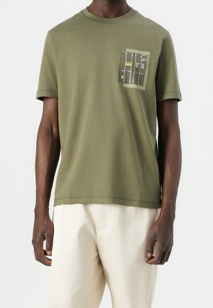 Homme portant un t-shirt à manches courtes vert olive avec un petit motif de conteneur sur la zone de la poche poitrine gauche et un pantalon blanc.