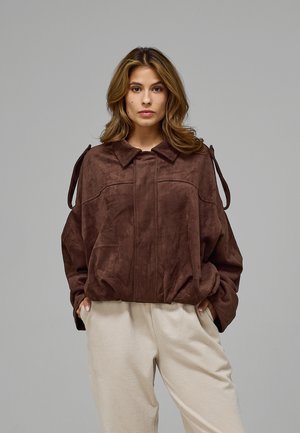Femme aux cheveux mi-longs porte une veste marron ample avec des liens aux manches et un pantalon beige clair texturé, debout devant un fond uni.