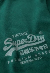 Grøn sweatshirt med et vintage SuperDry-logo i lysegrå. Stoffet har en blød tekstur, og teksten inkluderer både engelsk og japansk.