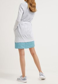 Cardigan a maglia grigio chiaro con cintura in vita, indossato su una gonna teal, abbinato a sneakers grigie e bianche. Tessuto texturizzato con maniche lunghe.
