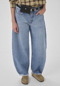 Lyseblå denim jeans med vide ben og flossede sømme, kombineret med et sort bælte med en lille lomme og beige ruskindssko uden snørre.