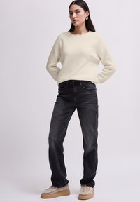 Vrouw staat met handen op de heupen, gekleed in een crèmekleurige ribgebreide trui, zwarte jeans, beige met bont gevoerde schoenen en gouden oorbellen op een witte achtergrond.