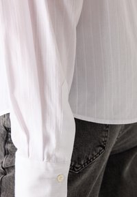 Hvit langermet skjorte med vertikale striper, med tettsittende mansjetter festet med en knapp, sammen med mørkegrå jeans.