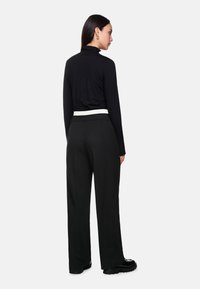 Woman facing sideways wearing a black turtleneck, high-waisted black wide-leg pants, white belt, and black chunky shoes on a white background.