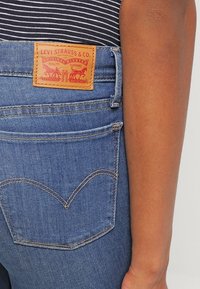 Detailní záběr na zadní kapsu modrých džínů s koženou etiketou Levi Strauss & Co a paží osoby viditelnou na pravé straně.