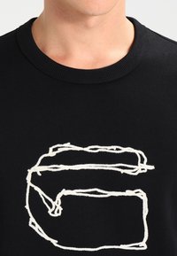 Černá mikina se kulatým výstřihem, která má bílou, vyšitou grafiku připomínající stylizované písmeno 'G'. Měkká tkanina s minimální texturou.