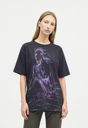 Femme aux longs cheveux blonds portant un T-shirt noir oversize avec un croquis violet d’un cheval au galop sur le devant.