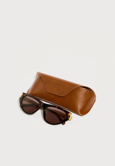 Γυαλιά ηλίου σε σχήμα γάτας με σχέδιο χελωνόφιδου και σκούρους φακούς δίπλα σε καφέ δερμάτινη θήκη με ανάγλυφο το όνομα της μάρκας, τοποθετημένα σε λευκή επιφάνεια.