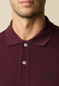 Burgundské polo tričko s texturovaným tkaním, s límečkem a tří knoflíkovou légou, a malým vyšitým logem na hrudi.