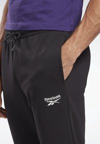 Black sweatpants with side pockets, drawstring waist, and white Reebok logo. Fabric appears soft with a smooth texture.