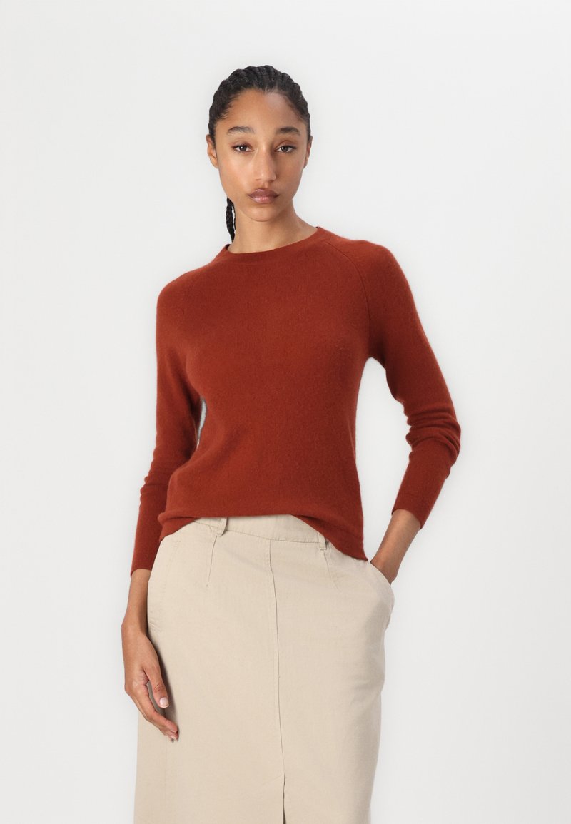 Rdzawoskrzydły sweter z miękkiego materiału, z okrągłym dekoltem i długimi rękawami, w połączeniu z jasnobeżową spódnicą.