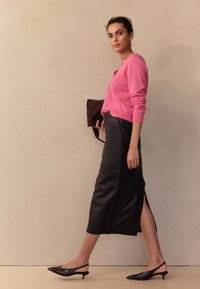 Różowy kardigan z dzianiny, czarna skórzana midi spódnica z bocznym rozcięciem oraz czarne czółenka na obcasie z paskiem z tyłu. Modelka trzyma brązową torebkę.