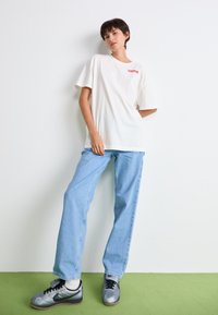 Př oversized bílém bavlněném tričku s červeným logem, spárovaném s světle modrými rovnými džínami a stříbrnými teniskami Nike na zelené podlaze.