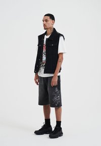 Gilet di jeans nero su una t-shirt bianca con grafica, abbinato a pantaloni corti grigi con una stampa illustrata. Sneakers nere e calzini alti completano il look.