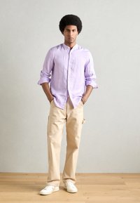 Lichtpaarse overhemd met lange mouwen en knopen, beige cargobroek, witte sneakers; model staat met handen in de zakken op een houten vloer.