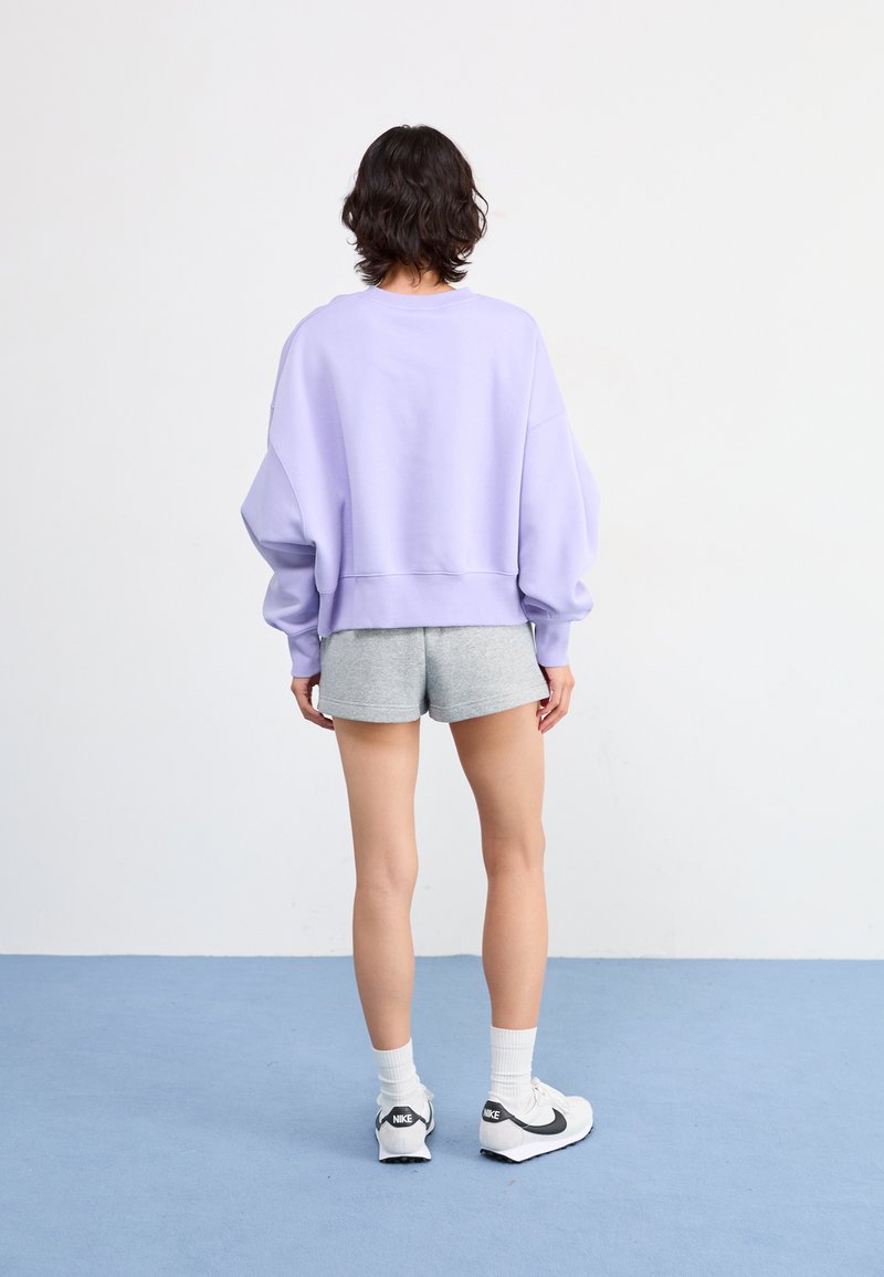 Fioletowa, oversize'owa bluza wykonana z miękkiego materiału, zestawiona z szarymi szortami. Ma okrągły dekolt i ściągacz na dolnym brzegu. Całość dopełniają białe buty sportowe.
