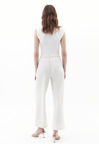 Witte mouwloze top gecombineerd met lichtgrijze, hoge taille, wijdvallende broek. Glad stoftextuur; model draagt gouden sandaaltjes met bandjes.