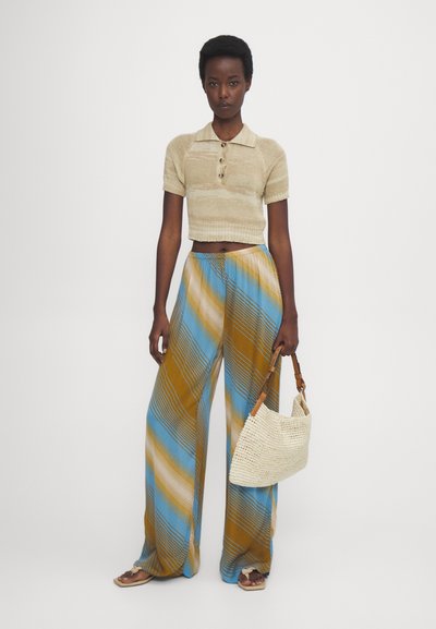 Beige cropped polo shirt met knoopdetails, gecombineerd met brede broeken met blauwe, gouden en bruine diagonale strepen, en een geweven tas.