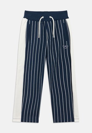 Granatowe i białe spodnie dresowe w paski z elastycznym, prążkowanym pasem, sznurkiem do regulacji i kieszeniami po bokach. Na lewej udzie znajduje się wyhaftowane logo.