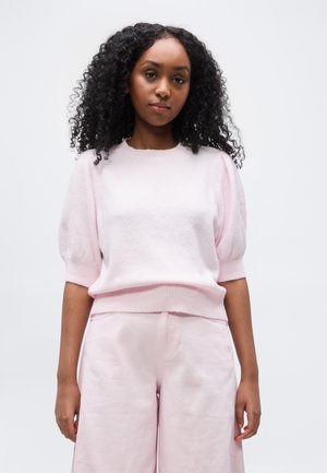Kvinne med krøllete svart hår iført en lys rosa kortermet genser og matchende bukser med vide ben, stående mot en ensfarget bakgrunn.