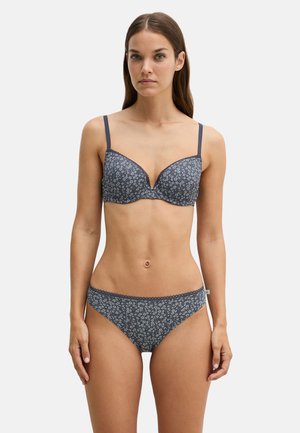 Vrouw draagt bijpassende donkerblauwe beha en slip met klein wit bloemenpatroon, staand tegen een effen lichte achtergrond.