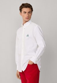 Joven con cabello castaño claro que lleva una camisa blanca de botones con un logo bordado y pantalones rojos, de pie con una mano en el bolsillo.