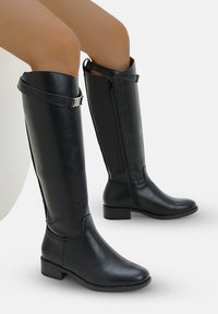 Bottes en cuir noires à hauteur de genou avec fermeture éclair latérale et sangle à boucle en haut, portées par une personne aux jambes nues sur un fond uni.