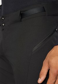 Juodos tamprios medžiagos kelnės su dviem mygtukų užsegimais, diržo kilpa ir užtrauktuku užsegama šonine kišenė su tekstūruotu paviršiumi.