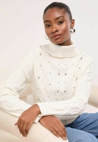 Fehér kötött pulóver magas gallérral, gyémántmintákkal és díszítő strasszokkal. Puha tapintású, bordázott ujjvégű.