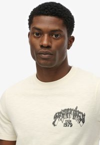 Cream-colored cotton t-shirt featuring a black graphic print with "Superdry Tour 1979." Classic crew neckline and short sleeves.
