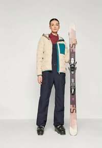 Giacca puffer beige con dettagli in teal, strato interno marrone, salopette scura e stivali da sci neri. Tiene un sci decorato con attacchi.