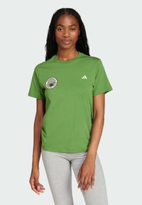 Woman with braided hair wearing a green t-shirt with a circular logo and a small triangular emblem, paired with light gray leggings.