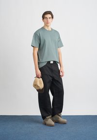 Hellgrünes Baumwoll-T-Shirt, schwarze, locker sitzende Hose, beige Baseballkappe und beigefarbene Stiefel mit strukturierten Sohlen. Schlichtes Design, minimale Akzente.