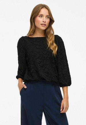 Blouse noire à texture avec un décolleté carré et des manches bouffantes, associée à un pantalon bleu marine, comportant des poches latérales.