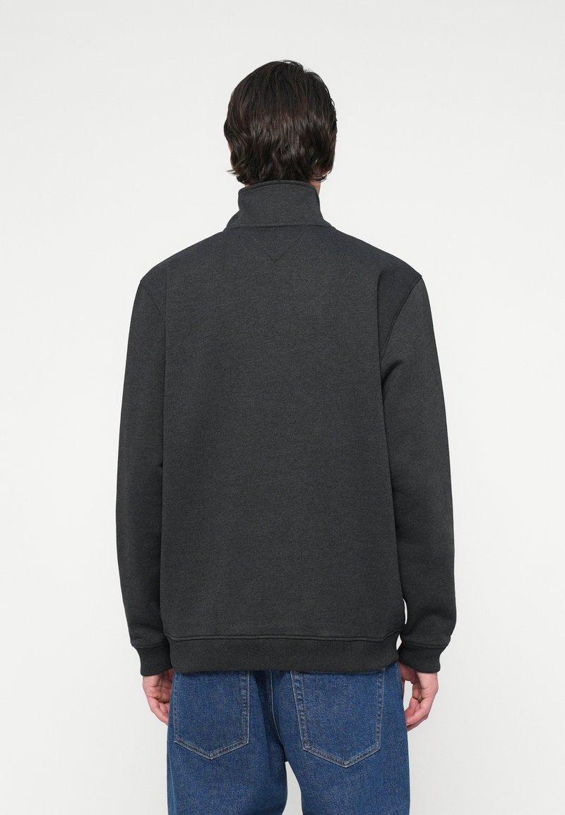 Mann trägt einen dunkelgrauen hochgeknöpften Pullover und blaue Jeans, von hinten vor einem schlichten weißen Hintergrund abgebildet.