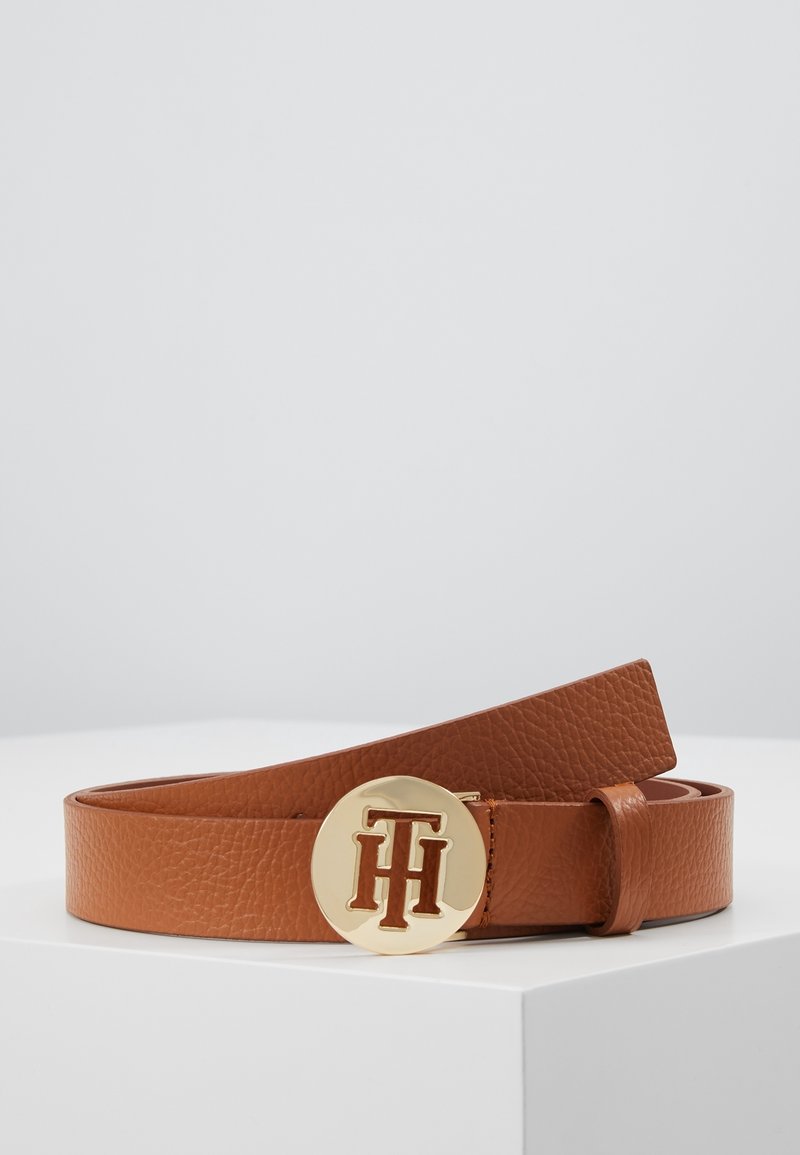Ceinture en cuir marron texturé enroulée sur une surface blanche avec une boucle ronde en or ornée du logo TH entrelacé.