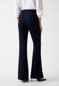 Jeans scuri in denim a zampa con vita alta, caratterizzati da due tasche posteriori e un piccolo dettaglio con logo sulla tasca destra. Texture liscia.