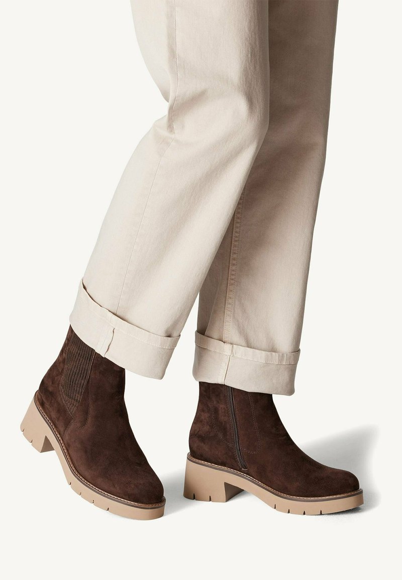 Brown suede ankle boots with a block heel, textured side panels, and a contrasting light sole. Worn with rolled-up cream-colored trousers.
