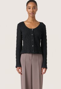 Cardigan nero texturizzato con scollo a barca, maniche lunghe e chiusura a bottoni, realizzato in un tessuto stropicciato e con una silhouette aderente.