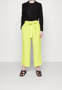 Czarna marynarka o skróconym kroju na czarnym topie, zestawiona z wysokim stanem, szerokimi neonowożółtymi spodniami z paskiem do związania. Czarne sandały na paski.