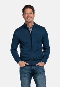 Blauwe ritska cardigan gemaakt van een geweven breimateriaal, met een hoge kraag en geribbelde manchetten, gecombineerd met een shirt met een patroon eronder.