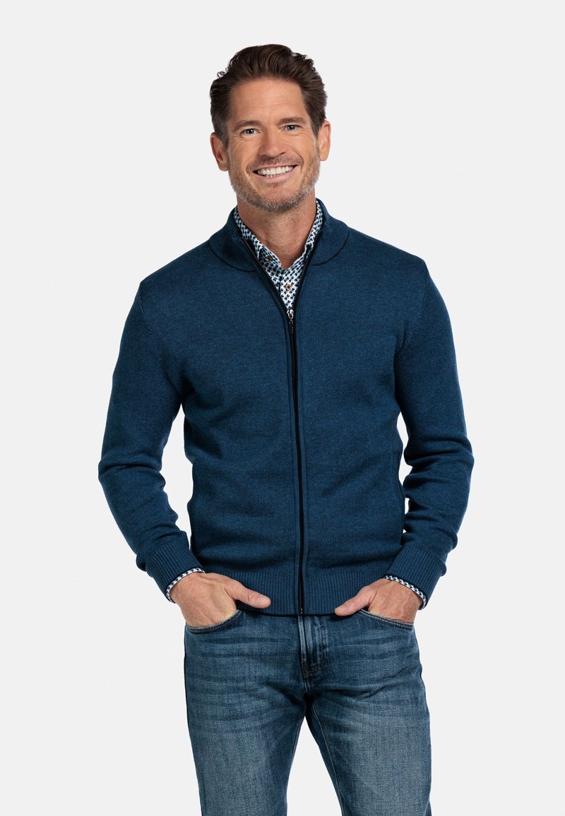 Blauwe ritska cardigan gemaakt van een geweven breimateriaal, met een hoge kraag en geribbelde manchetten, gecombineerd met een shirt met een patroon eronder.