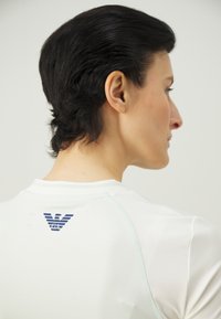 Bela atletska majica z logotipom na zgornjem delu hrbta, zaobljenega ovratnika in subtilnim kontrastnim šivanjem ob šivih.