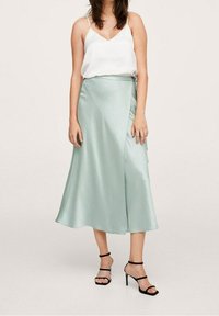 Una gonna midi in satin verde chiaro con design avvolgente, caratterizzata da un dettaglio con fiocco laterale. Abbinata a un top bianco e a sandali neri con cinturini.
