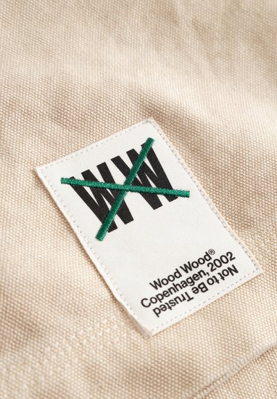 Zbliżenie na beżową tkaninę z białą prostokątną metką, na której widnieją czarne litery „W W” przecięte zielonym, haftowanym symbolem X oraz tekst „Wood Wood Copenhagen 2002.”