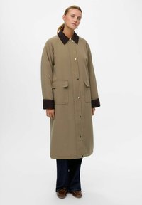 Lang beige frakke med en mørkebrun krave, knaplukning, to frontlommer og en afslappet pasform. Båret over mørke jeans og sko.