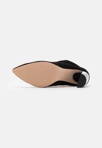 Zapato de tacón alto de ante negro con punta afilada, suela de goma beige y un acento plateado en el tacón. Superficie texturizada y diseño elegante.