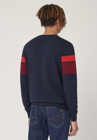 Maglione blu navy con accenti a righe rosse e marroni sulle maniche. Polsini e orlo a costine, texture morbida, design classico con scollatura a girocollo.